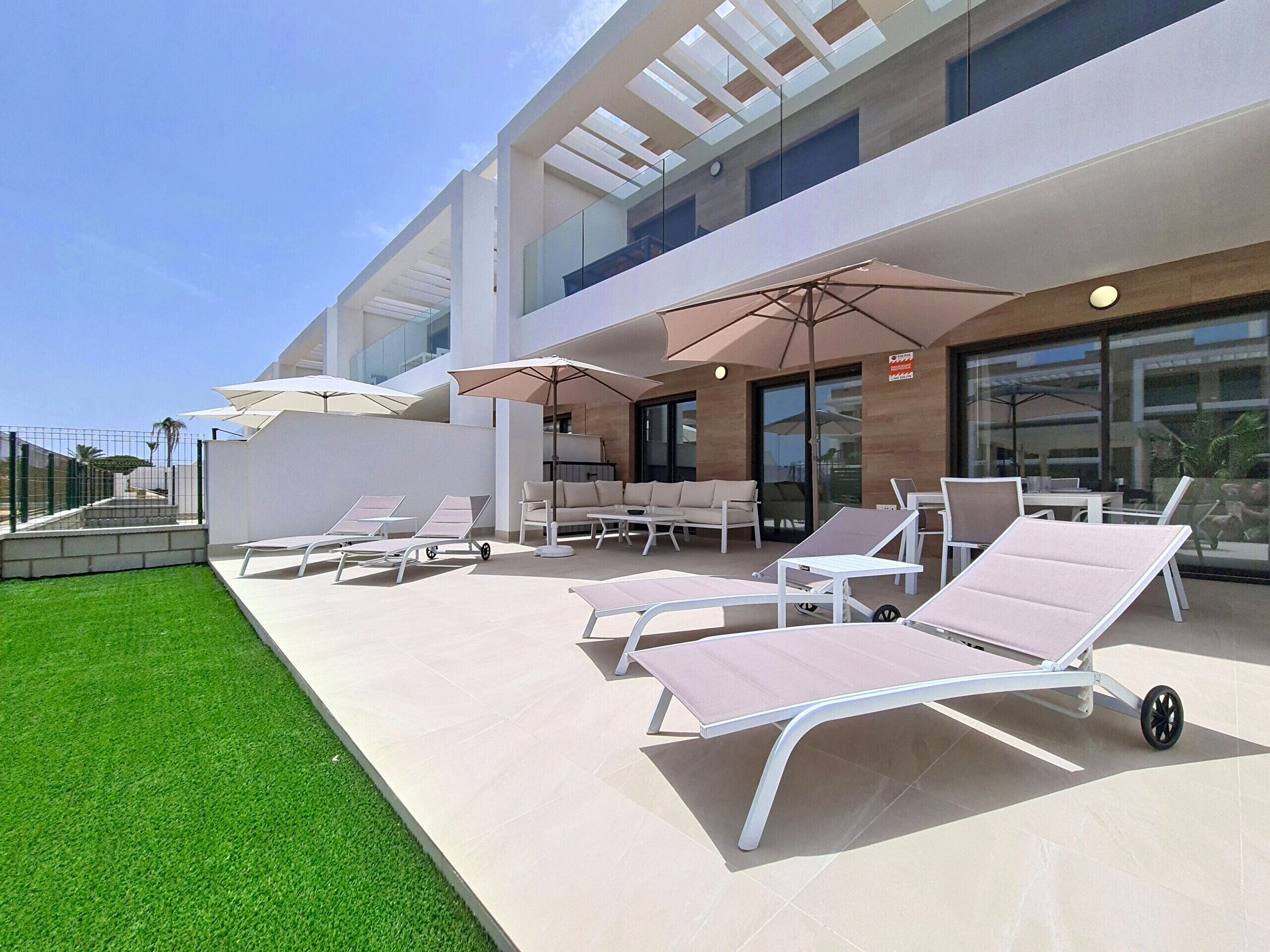 A modern outdoor lounge area with sun loungers and umbrellas, a glass-enclosed building in the background, and a well-maintained lawn.