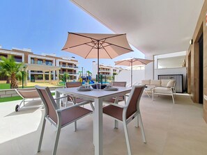 A patio area with a table and chairs, umbrellas, and a view of a pool and buildings in the background. - Ground Floor Oasis at La Serena Golf (Los Alcazares)