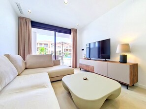A modern hotel room with a sofa, a flat-screen TV, a lamp, and a view of the outdoor pool area. - Ground Floor Oasis at La Serena Golf (Los Alcazares)