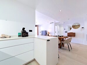 Een moderne keuken met een koffiezetapparaat, een eethoek met een tafel en stoelen, en een woonruimte met een televisie en een spiegel.