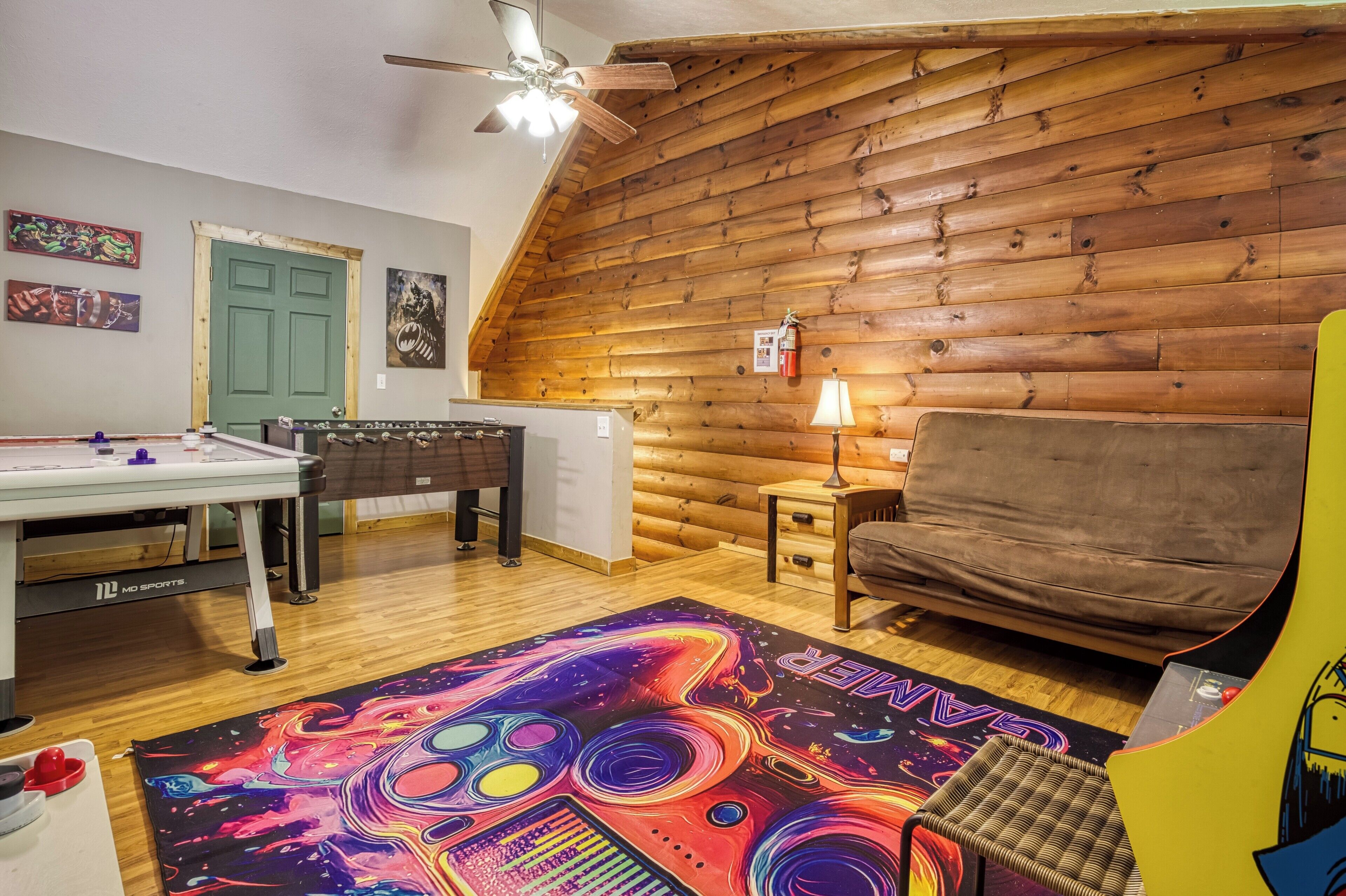 A room with a wooden sloped ceiling, a futsal table, a sofa, a colorful rug, and a lamp.