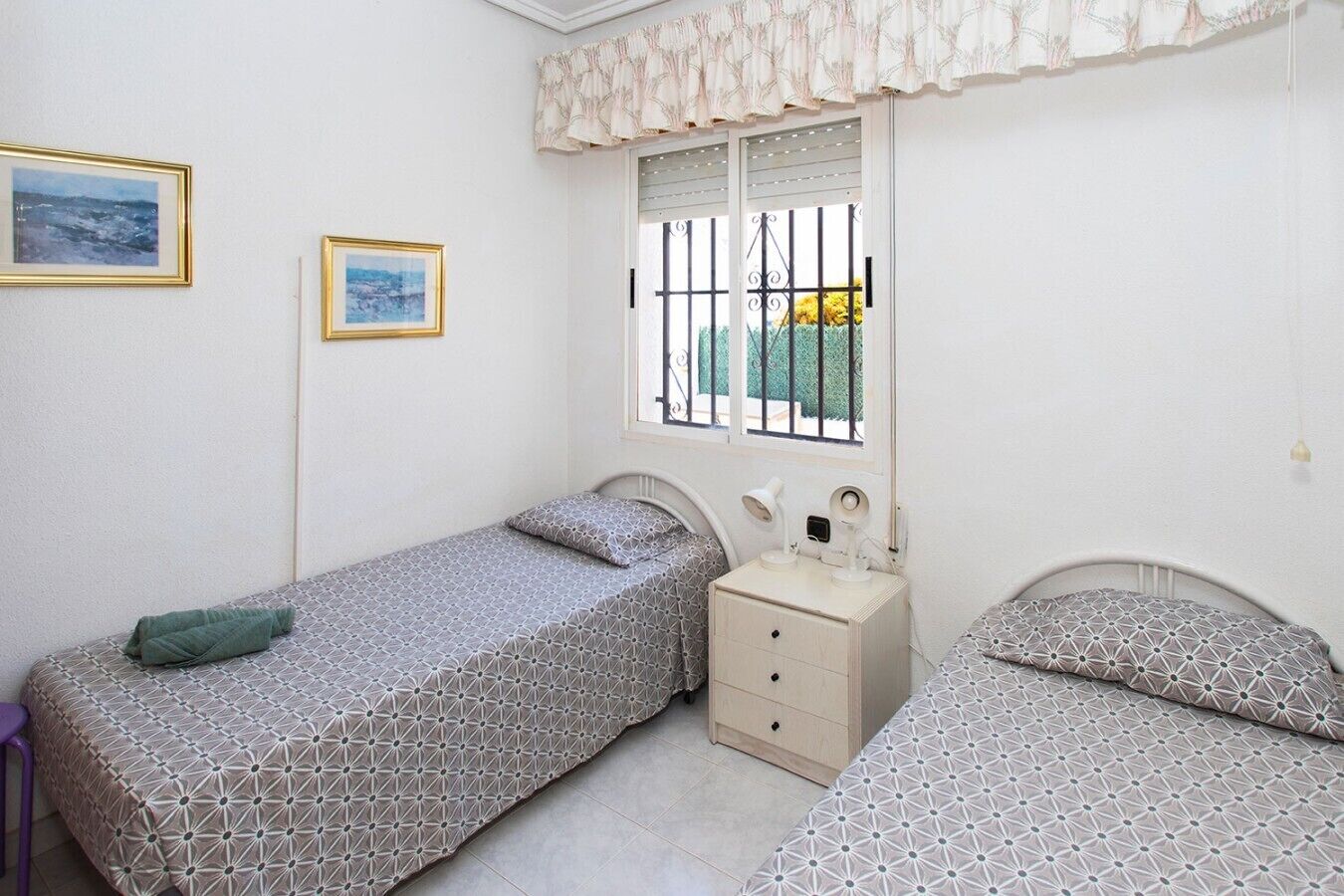 Ein Schlafzimmer mit zwei Betten, einem Nachttisch, einem Fenster mit Gitterstäben und zwei gerahmten Bildern an der Wand.