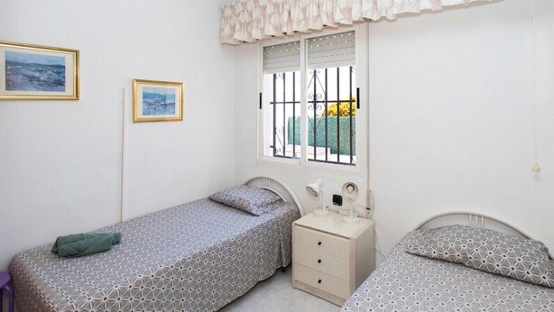 Une chambre avec deux lits, une table de chevet, une fenêtre avec des barreaux et deux tableaux encadrés accrochés au mur.