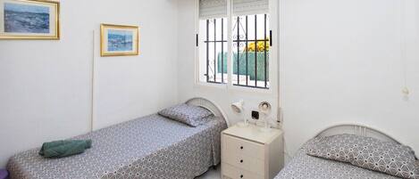 Ein Schlafzimmer mit zwei Betten, einem Nachttisch, einem Fenster mit Gitterstäben und zwei gerahmten Bildern an der Wand.