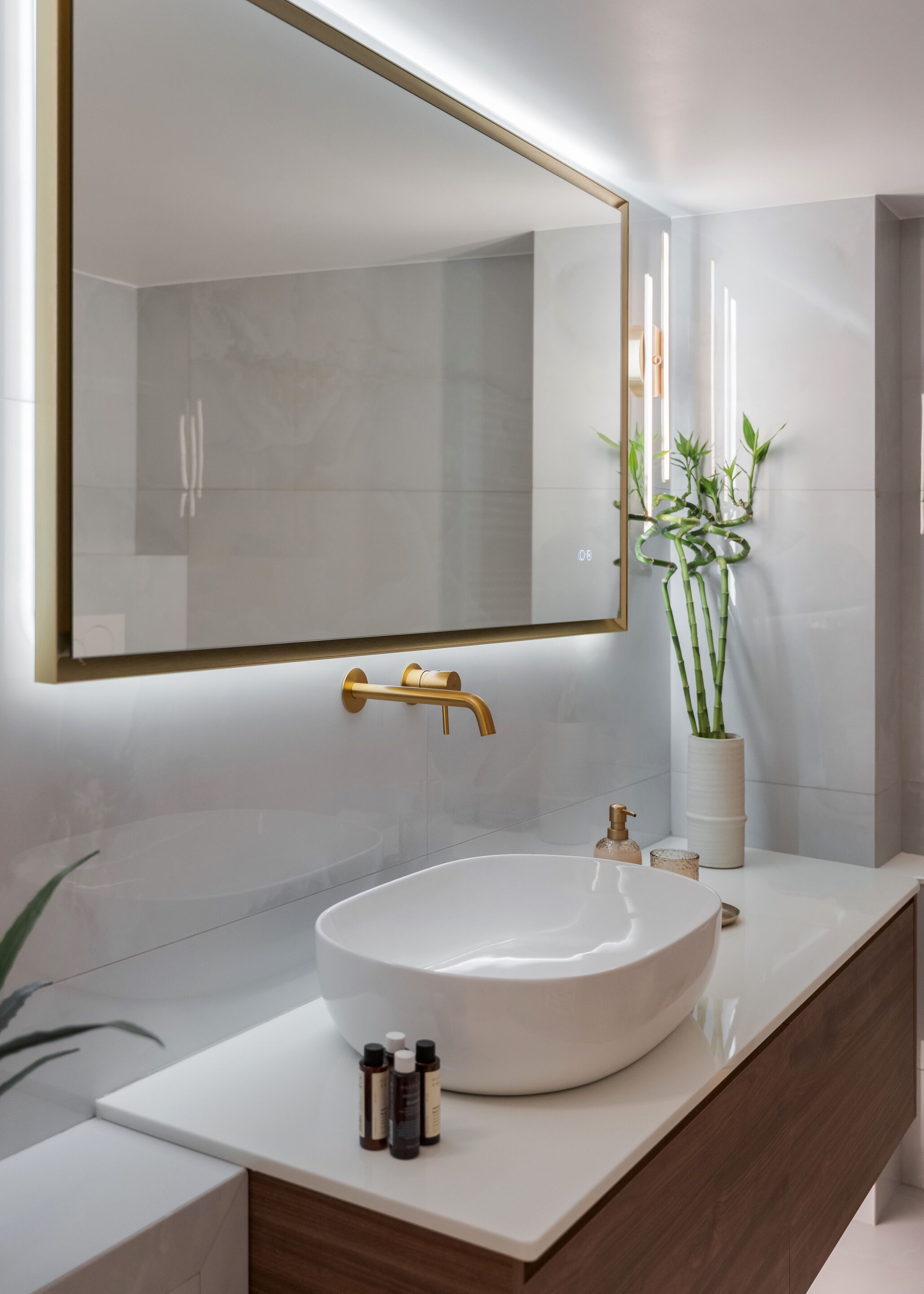 Un baño moderno con un espejo grande, un grifo dorado, un lavamanos blanco y una planta de bambú en maceta.