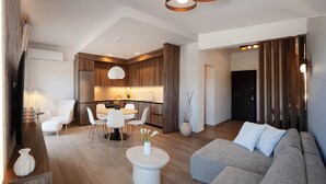 Una sala de estar moderna con un sofá gris, un comedor con una mesa y sillas blancas, y una cocina con armarios de madera y un horno empotrado.