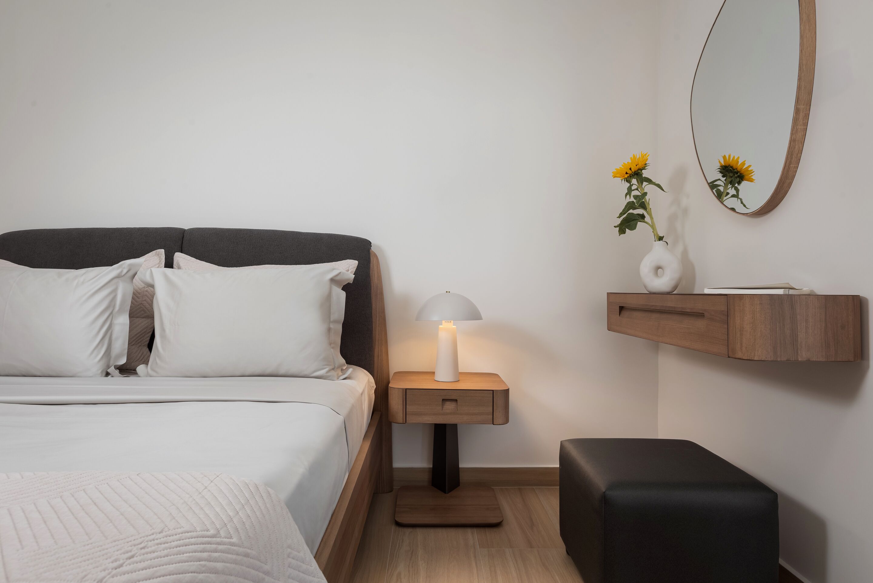 Un dormitorio ordenado con cama, mesita de noche con lámpara, un pequeño banco de madera y estante en la pared con un florero.