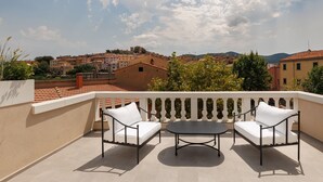 Un balcon avec deux chaises blanches dotées de coussins et une petite table ronde, d’où l’on peut admirer une ville aux toits de tuiles et un ciel dégagé.
