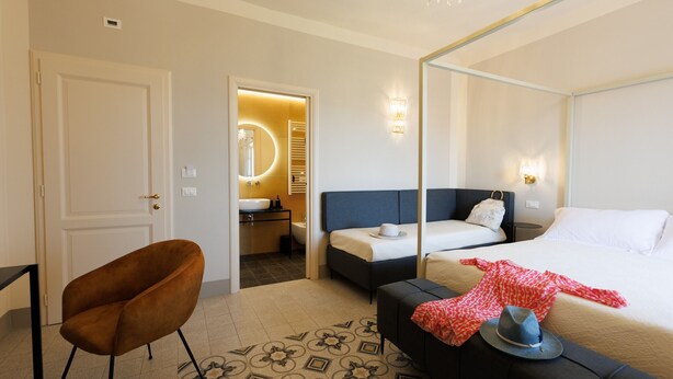 Une chambre d’hôtel avec un lit, un canapé, une chaise, une salle de bain avec un miroir et un lustre.