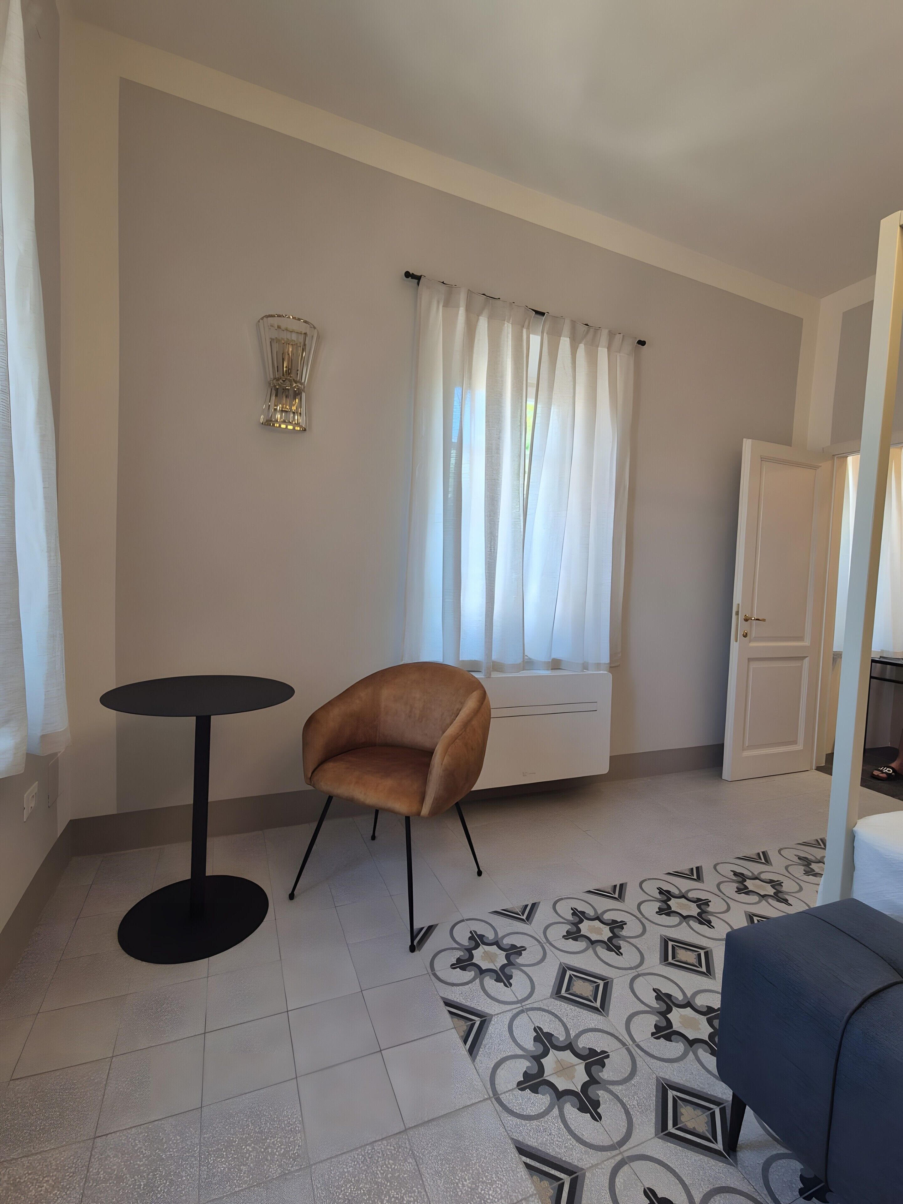 Ein Zimmer mit einem einzelnen braunen Stuhl, einem kleinen runden Tisch, einem gemusterten Boden, einem Fenster mit Vorhängen und einer Tür.