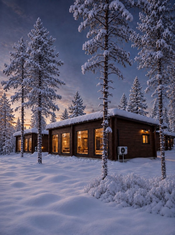 A wooden cabin with large windows, surrounded by snow-covered trees, set in a snowy landscape.