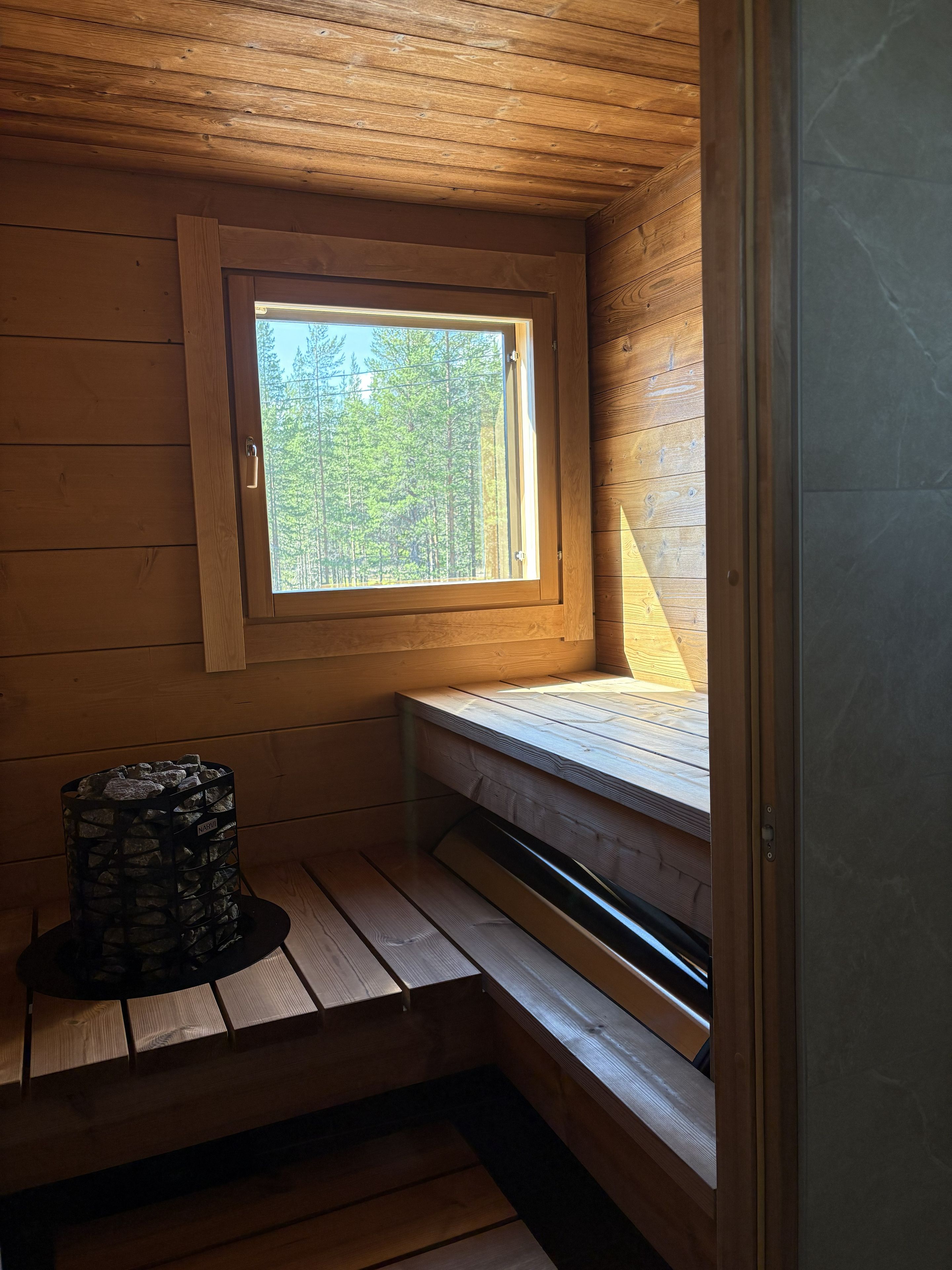L’intérieur d’un chalet en bois, avec un plafond en pente, une fenêtre donnant sur les arbres, un banc intégré et une cheminée en pierre.