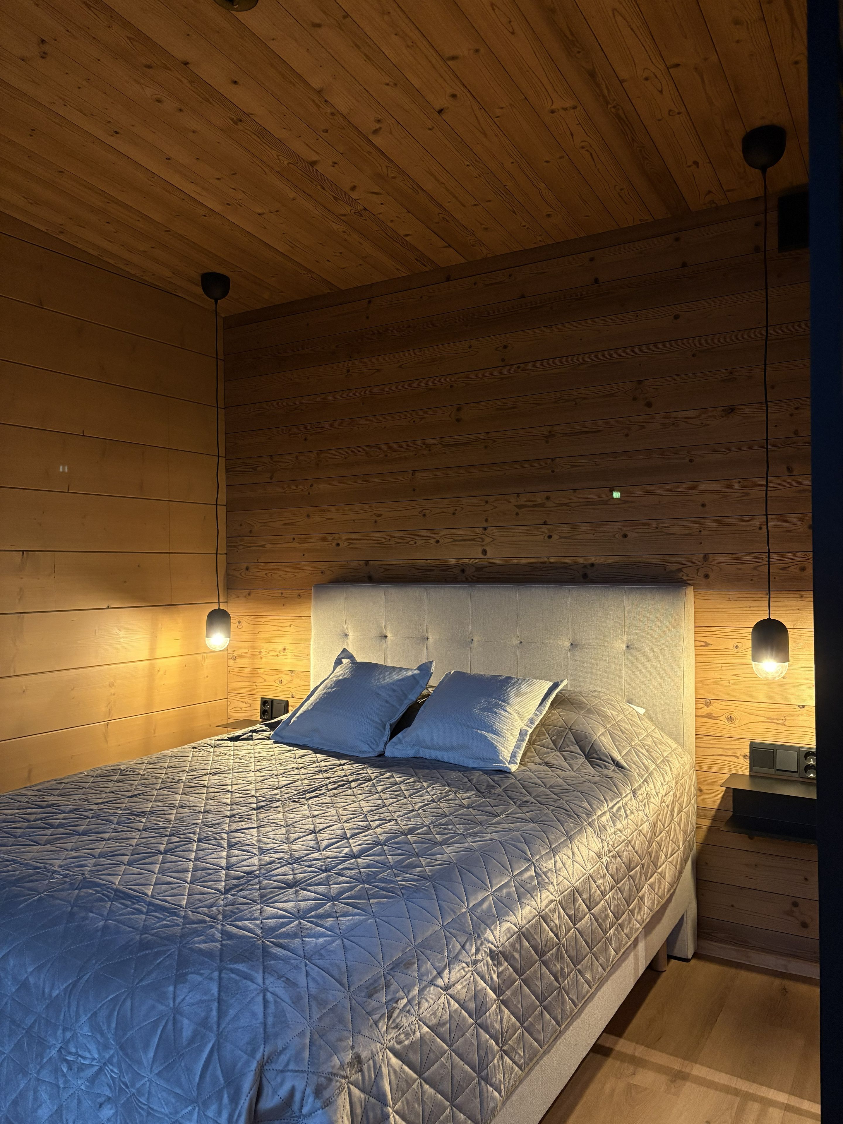 Une chambre avec un plafond en bois, un lit recouvert d’une courtepointe matelassée, deux oreillers, une table de chevet avec une lampe et un luminaire fixé au mur.