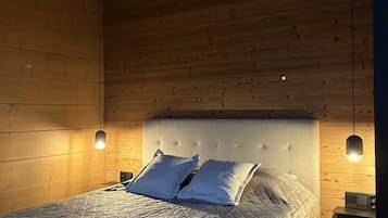 Une chambre avec un plafond en bois, un lit recouvert d’une courtepointe matelassée, deux oreillers, une table de chevet avec une lampe et un luminaire fixé au mur.