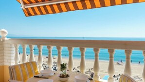 A balcony with a table set for dining, overlooking a beach with umbrellas and people. - Ocean Views on the Sabinillas Boardwalk (San Luis de Sabinillas)