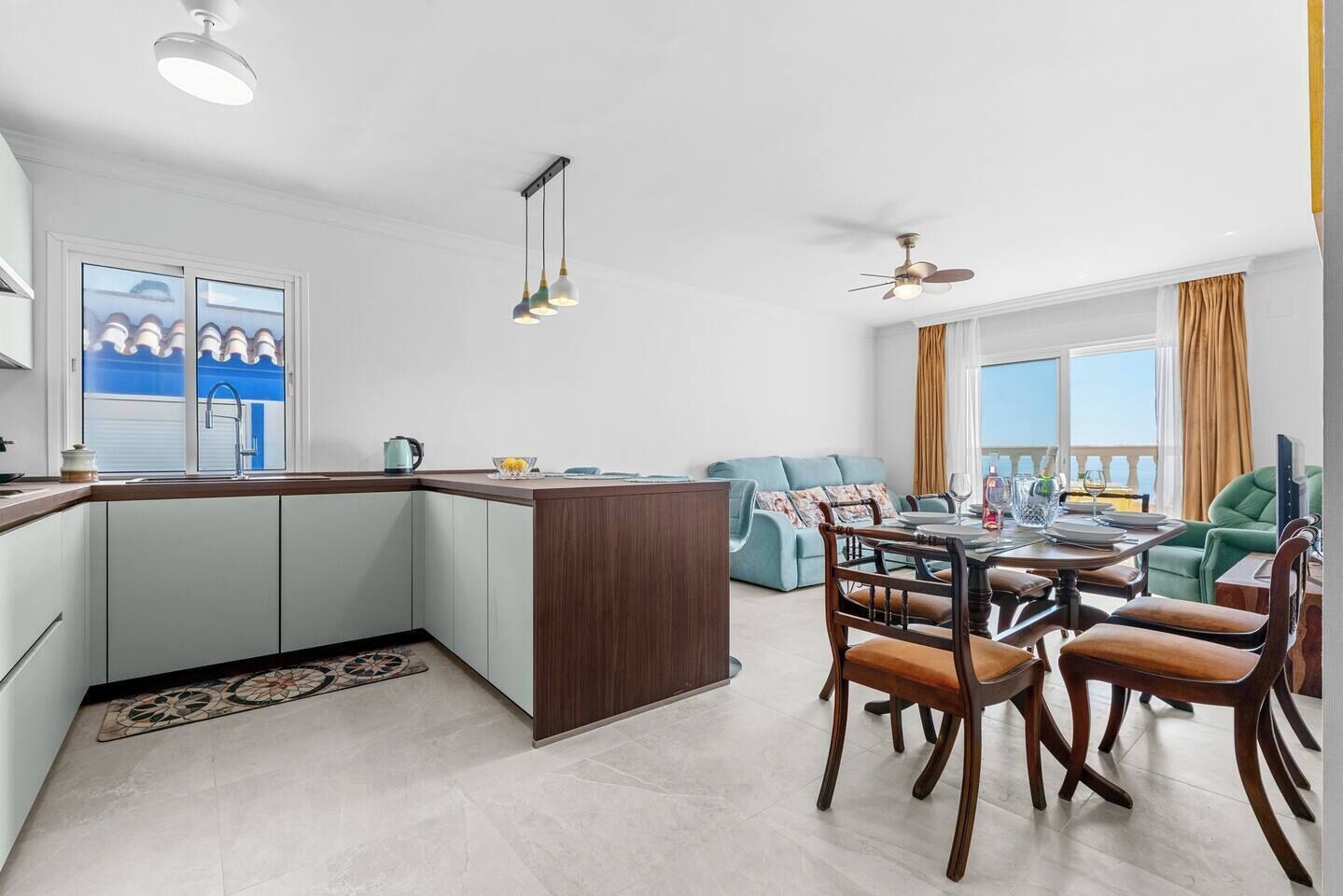 A modern kitchen with a central island, dining area with wooden chairs, and a view of the sea through large windows.