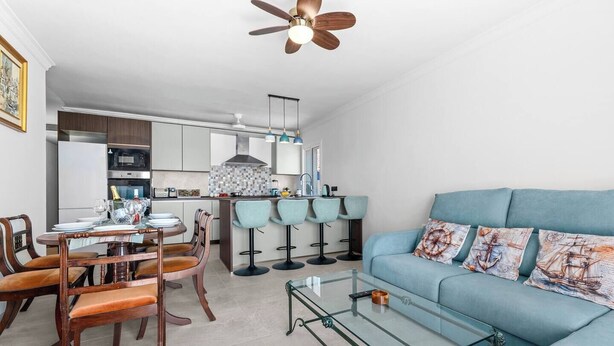 A modern living room with a blue sofa, a glass coffee table, and a dining area with wooden chairs and a table. - Ocean Views on the Sabinillas Boardwalk (San Luis de Sabinillas)