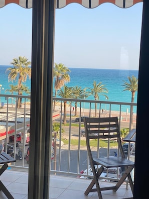 A balcony with a view of the sea, palm trees, and a parking area with cars and a building with signage. - Beautiful 2-bedroom apartment with seaview in gorgeous Nice - Azure Paradise (Nice)