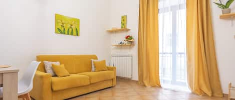A living room with a yellow sofa, a white chair, a radiator, and a window with yellow curtains.