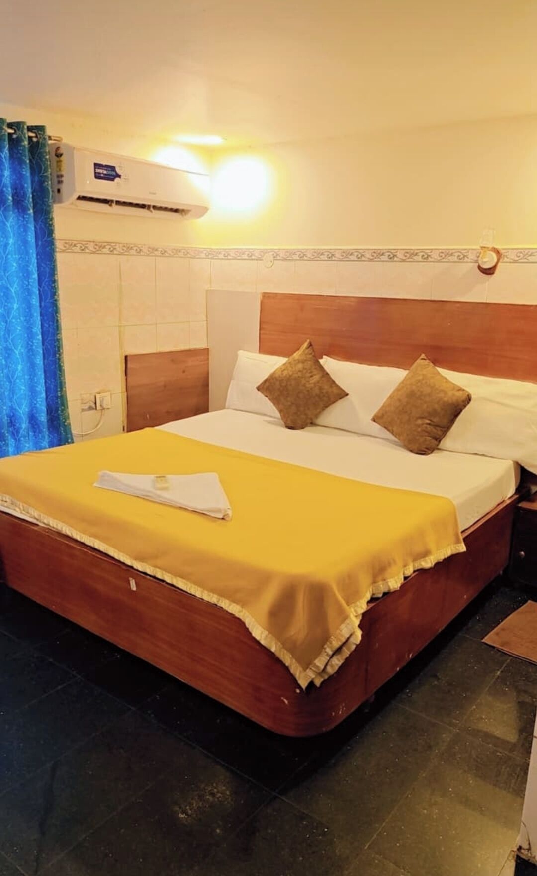Una habitación de hotel con cama, almohadas, mesita de noche, aire acondicionado y cortina.
