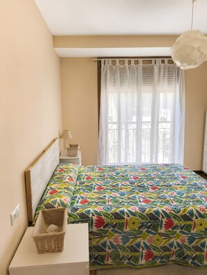 Une chambre à coucher comprenant un lit, une table de chevet, une lampe, une fenêtre avec des rideaux légers et un luminaire suspendu.