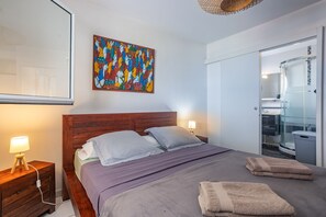 A hotel room with a bed, a nightstand, a lamp, a bathroom with a glass shower enclosure, and a colorful abstract painting on the wall. - Ti Bato: Apartment with ocean views and the beach within walking distance (Les Trois-Ilets)