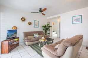 A living room with a sofa, a coffee table, a TV on a stand, a ceiling fan, and a large plant. - Ti Bato: Apartment with ocean views and the beach within walking distance (Les Trois-Ilets)