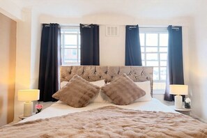 A hotel room with a large bed, two bedside lamps, a fluffy rug, and a window with curtains. - Mayfair Beautiful Family Apartment (London)