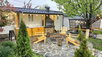 A patio area with a stone floor, wooden chairs, a fire pit, and a barbecue grill.
