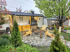 Een terras met stenen vloer, houten stoelen, een vuurkorf en een barbecue.