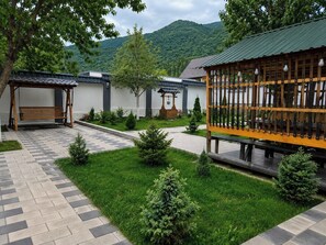 A courtyard with a wooden bench, a pergola with hanging lights, and a small garden area with neatly trimmed grass and shrubs.
