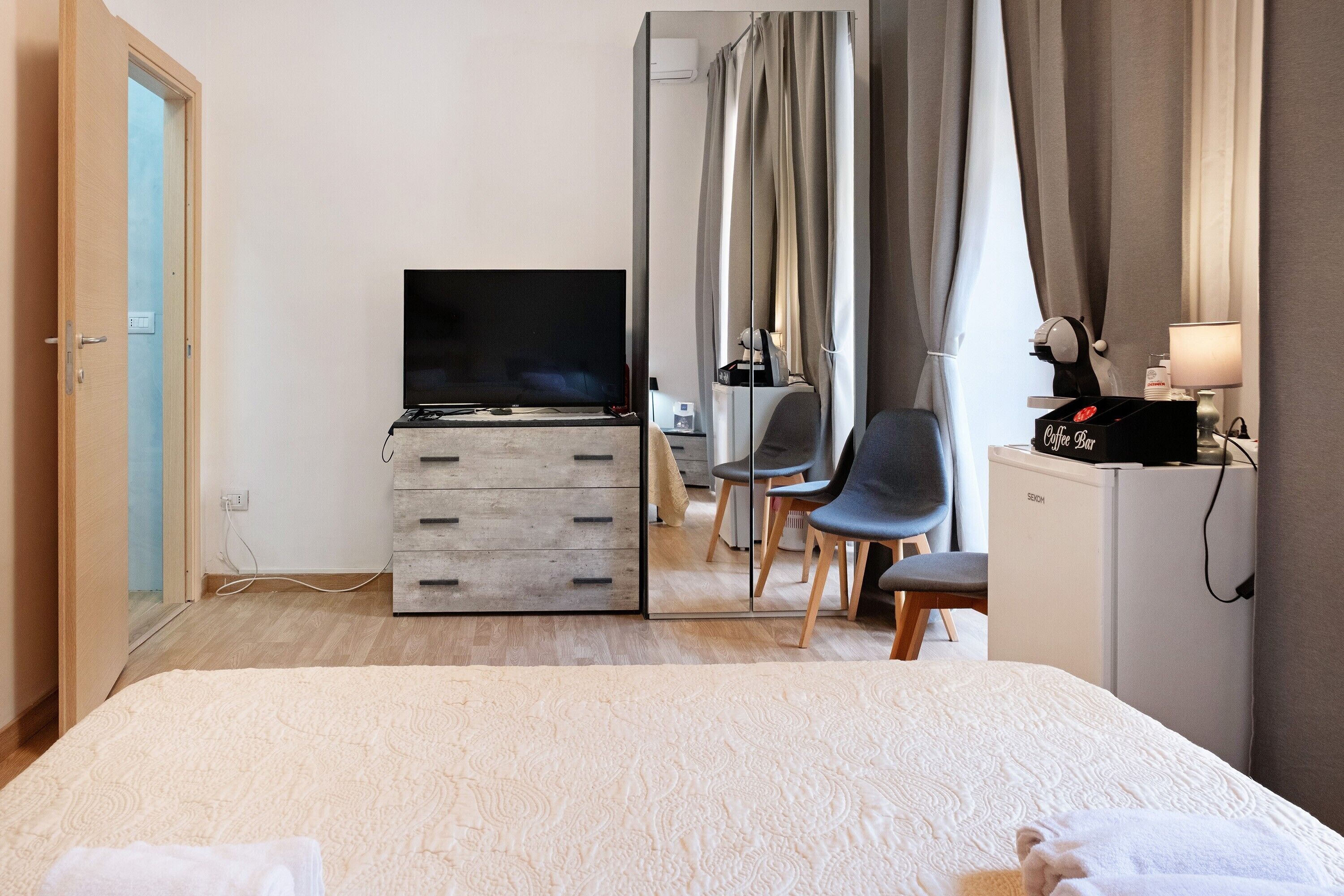 A hotel room with a bed, a TV, a desk with drawers, a chair, a coffee machine, and a mirror.