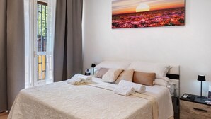A neatly made bed with a patterned bedspread, a bedside table with a lamp, and a painting of flowers on the wall. - Home Joséphine - Caserta - Triple Room with Private Bathroom (Caserta)