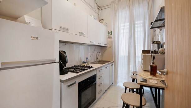 A modern kitchen with white cabinetry, a gas stove, a built-in oven, and a dining area with two stools. - Home Joséphine - Caserta - Triple Room with Private Bathroom (Caserta)