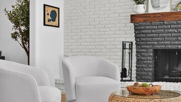 Una sala de estar moderna con una chimenea de ladrillo blanco, un sillón blanco y una mesa de centro de mimbre con un cuenco de frutas.