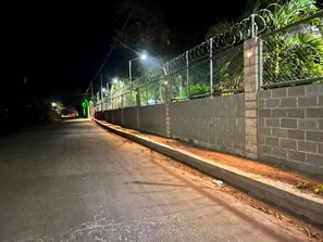 Ein beleuchtetes Straßenbild bei Nacht mit einer gepflasterten Straße, einer Ziegelmauer mit Stacheldraht und einem grünen Ampelsignal in der Ferne.