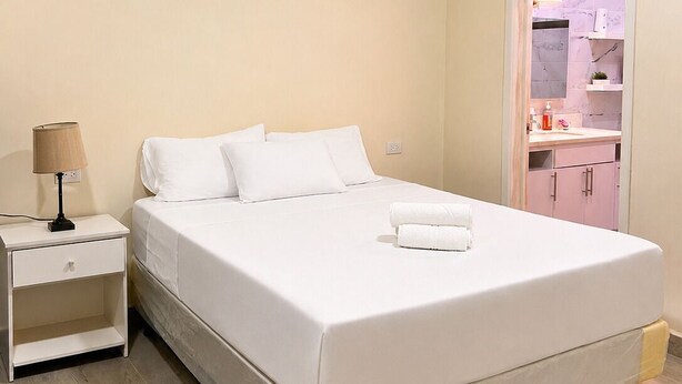 Habitación de hotel con una cama individual, una mesita de noche, una lámpara y vista al baño por una puerta abierta.