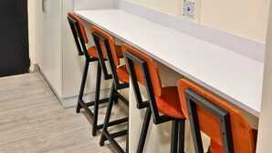 Una kitchenette con un refrigerador blanco, una encimera blanca y cuatro sillas naranjas con estructura negra.