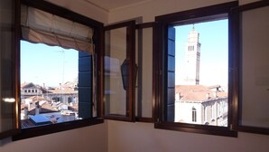 A room with two windows, one with a curtain and one with a shutter, overlooking a cityscape with buildings and a tower. - Panoramic view in San Marco overlooking the rooftops (Venezia)