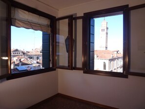 Ein Zimmer mit zwei Fenstern, eines mit Vorhang, das andere mit Rollo, mit Blick auf eine Stadtlandschaft mit Gebäuden und einem Turm.