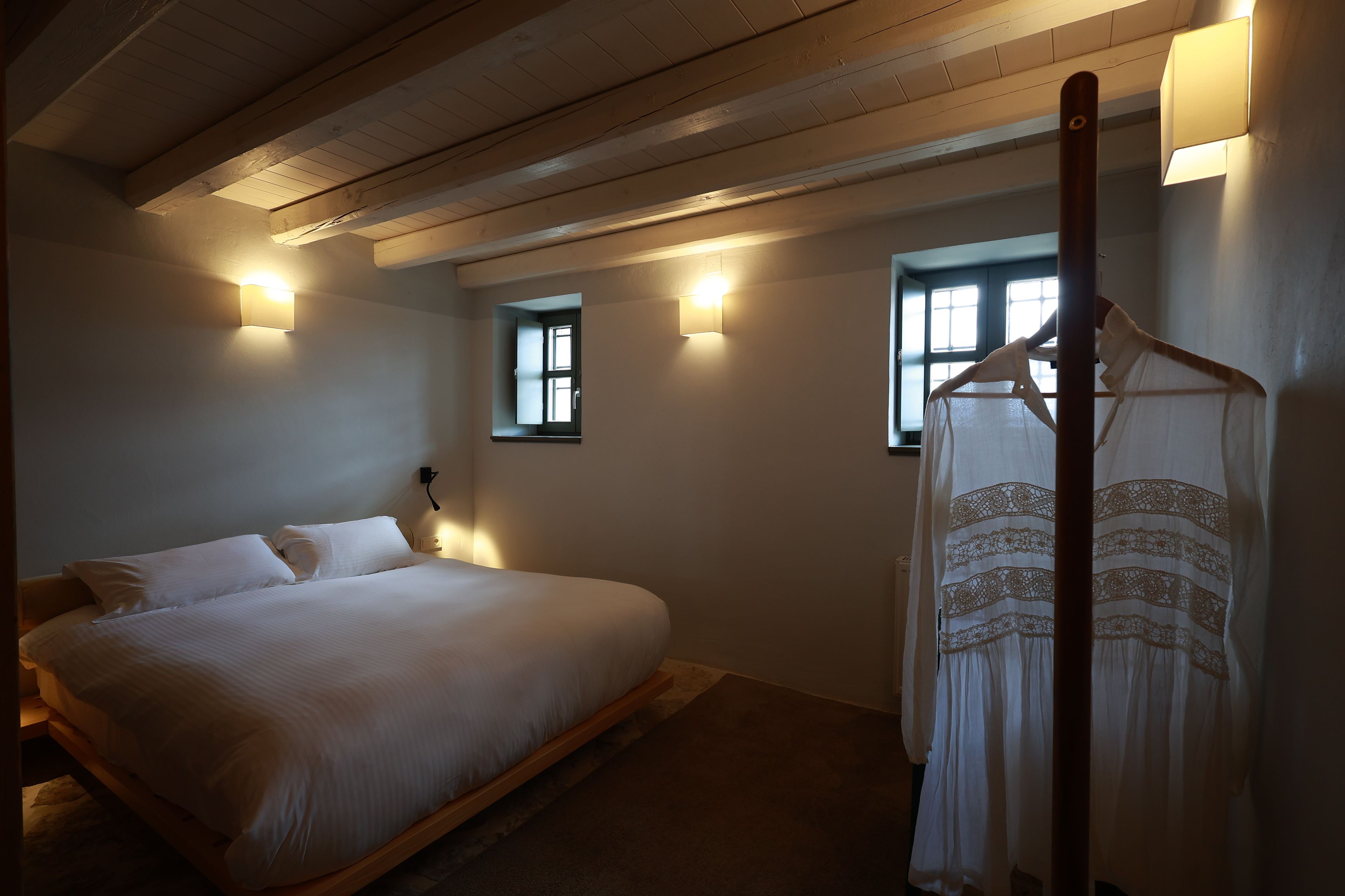Une chambre à coucher avec un lit, un vêtement suspendu et deux fenêtres équipées de lampes fixées au mur.