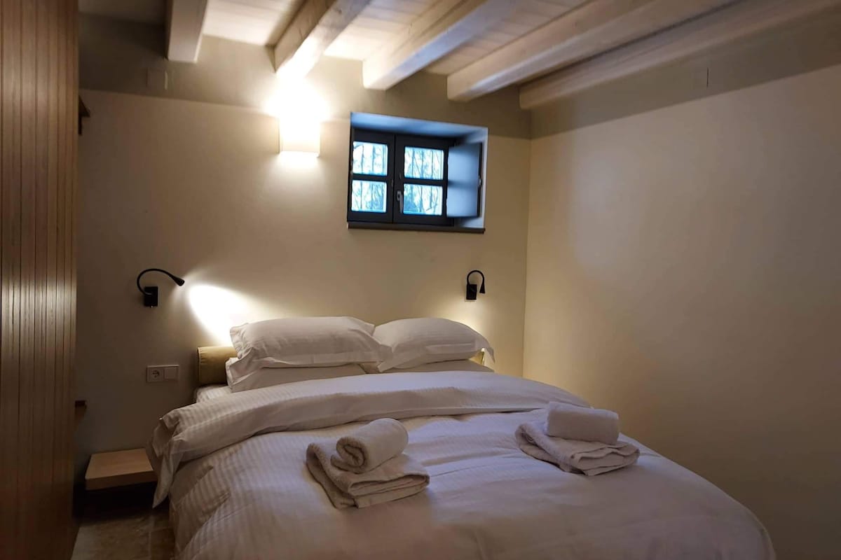 Une chambre d’hôtel avec un lit, deux lampes fixées au mur, une fenêtre avec des stores et un plafond en bois.