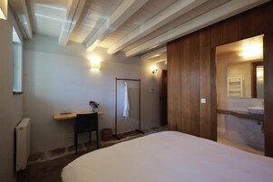 A bedroom with a bed, a desk, a chair, a wooden wardrobe, and a bathroom with a sink and mirror.