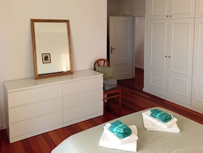 Ein Zimmer mit weißem Schminktisch, einem Spiegel mit goldener Rahmung, einem Stuhl mit grüner Sitzkissen, einer Tür und einem kleinen Tisch mit gefalteten Handtüchern.