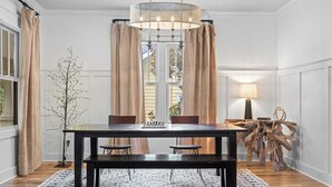 A dining area with a dark wooden table, two chairs, a bench, a chandelier, and a large window with curtains. - Above Nashville West End Rhythm | Music Studio | Premium Location | Luxe (Nashville)