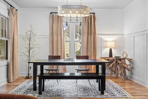 A dining area with a dark wooden table, two chairs, a bench, a chandelier, and a large window with curtains. - Above Nashville West End Rhythm | Music Studio | Premium Location | Luxe (Nashville)