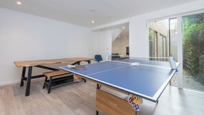 A ping pong table with a net, a blue chair, and a wooden bench in a room with white walls and a glass door leading outside. - Bright Modern 3BDR Family Home with Private Patio (Greater London)