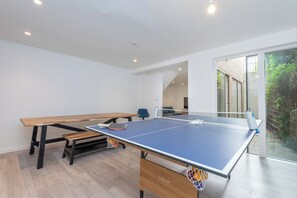 Ein Tischtennistisch mit Netz, ein blauer Stuhl und eine Holzbank in einem Raum mit weißen Wänden und einer Glastür, die nach draußen führt.