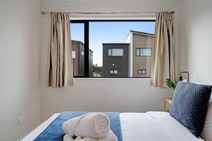 A hotel room with a bed, a window with curtains, a view of buildings outside, and a bedside table with a lamp.