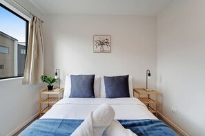 A hotel room with a bed, two bedside tables, a potted plant, a wall-mounted artwork, and a window with curtains.
